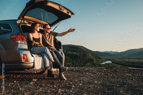 Young Traveler Couple on a Road Trip, Man and Woman Sitting on the Opened Trunk of Their Car Over Sunset