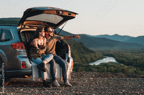 Young Traveler Couple on a Road Trip, Man and Woman Sitting on the Opened Trunk of Their Car Over Sunset