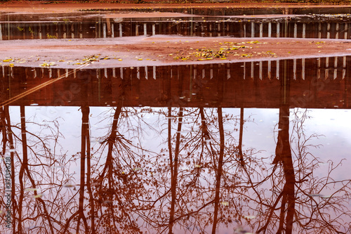 Autumn city background. Puddles on playground in rainy weather. Water on red clay tennis court and reflections of trees on it. Wet tennis court after rain. End of season for outdoor sports