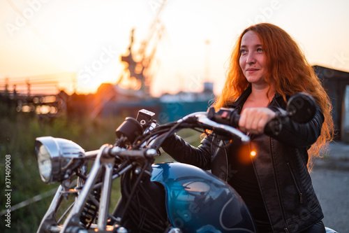 Curly red-haired woman in a black leather jacket sits on a motorcycle at sunset. Portrait of a serious girl driving a bike. © Михаил Решетников