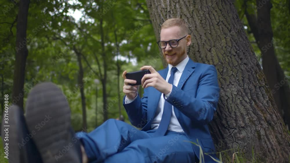 Cheerful businessman sitting under tree and celebrating winning video game on smartphone Cheerful businessman sitting under tree and celebrating winning video game on smartphone
