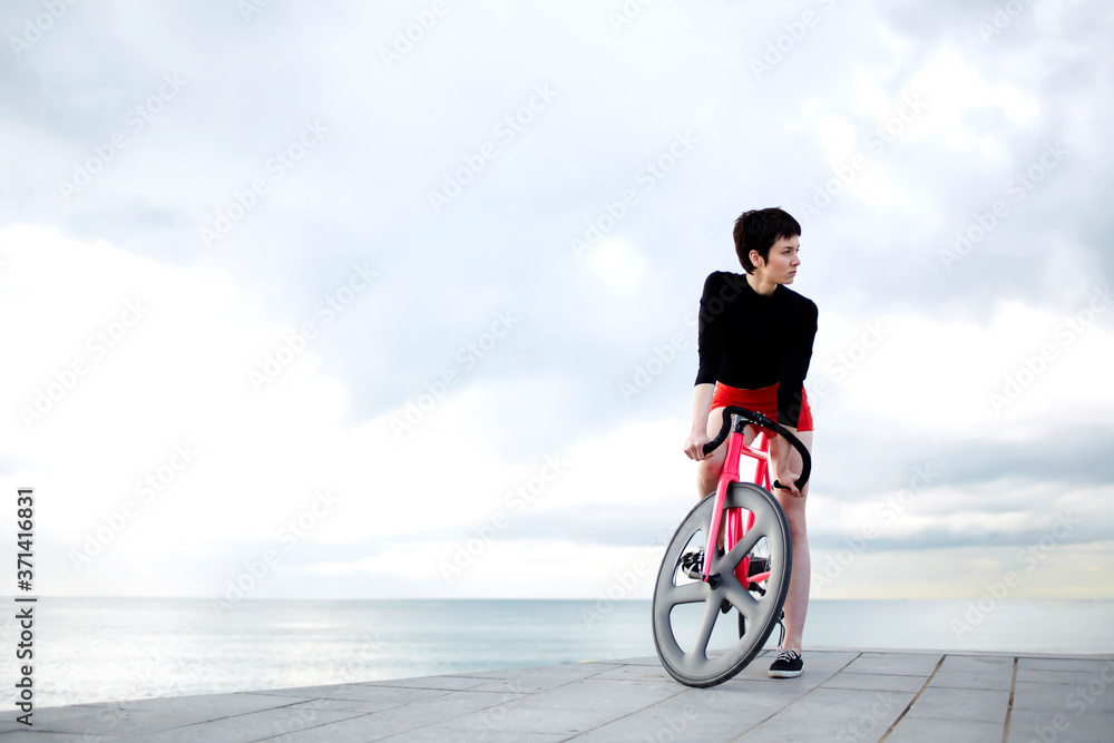 Fototapeta premium Portrait of stylish hipster woman sanding on concrete pier with her pink sport fixed gear bicycle against cloudy sky background with copy space area for your content, female cyclist resting after ride