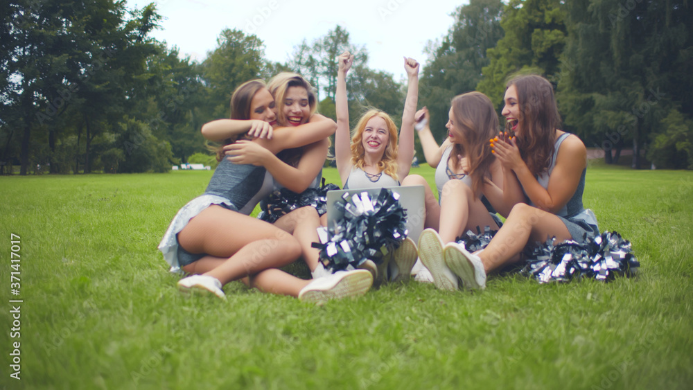 Cheerleaders sitting on grass in park and checking competition results on laptop celebrating victory Cheerleaders sitting on grass in park and checking competition results on laptop celebrating victory