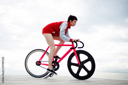 Fototapeta Naklejka Na Ścianę i Meble -  Full length portrait of an attractive hipster girl riding on her sport fixed gear bicycle along seashore on copy space cloudy sky background, female with rental bike enjoying weekend stroll outdoors