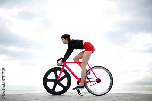 Fototapeta Naklejka Na Ścianę i Meble -  Full length portrait of professional female rider dressed in red sport shorts ride on bicycle along concrete pier seashore with copy space cloudy sky,young hipster girl enjoys recreation time outdoors