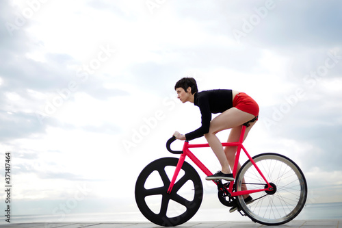 Full length portrait of young woman cyclist dressed in sexy red shorts riding bicycle along seashore against gray cloudy sky background with copy space area for your text message or advertise content