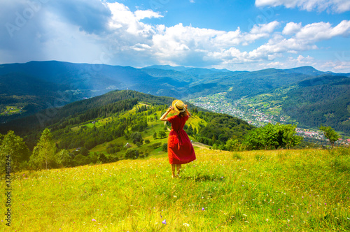 Beautiful girl in the mountains. Woman in linen dress and straw hat travelling. Amazing summer nature around. Harmony and wanderlust concept. Rustic natural style. Wind blowing for dynamic photo.
