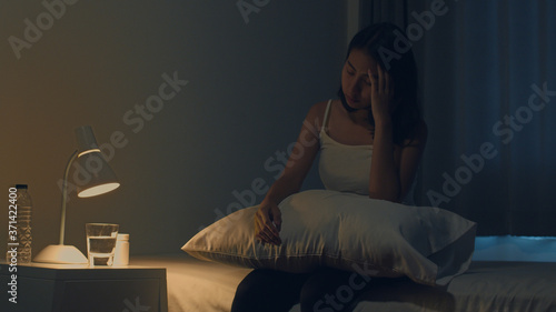 Beautiful Asian young woman sitting on bed take sleeping pill or night medicine in bedroom. Unhealthy sick Indian female suffers from insomnia or headache, depressed girl holds antidepressant meds.