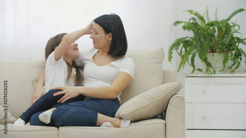 Beautiful daughter whispering in mother's ear, telling her a secret, while they are sitting on the sofa, in homely atmosphere, hugging. Beautiful daughter whispering in mother's ear, telling her a secret, while they are sitting on the sofa, in homely atmosphere, hugging.