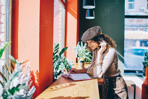 Trendy smiling female writer in stylish look creating new chapter for book standing at cozy cafeteria, positive hipster girl using notebook for course work spending free time at university campus © BullRun