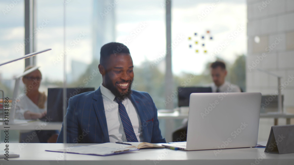 Fototapeta premium Young african businessman smiling celebrating success while looking at laptop screen in office