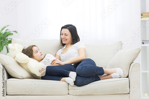 Charming little girl and her beautiful young mom smiling while sitting on sofa at home.