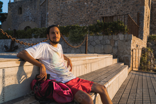 Young spanish man with white shirt and red shorts sitting on a stairs at a viewpoint at the village of Soller (Serra de Tramuntana, Mallorca, Spain) during the golden hour