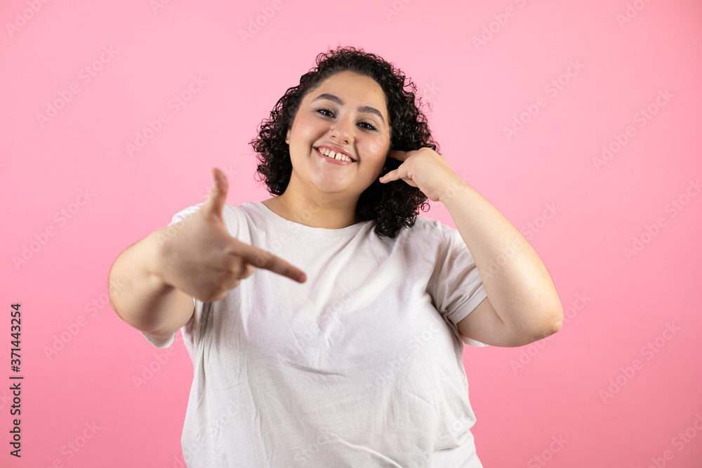 Young beautiful woman doing the “call me” gesture with her hands. Wearing white t-shirts on pink background Young beautiful woman doing the “call me” gesture with her hands. Wearing white t-shirts on pink background