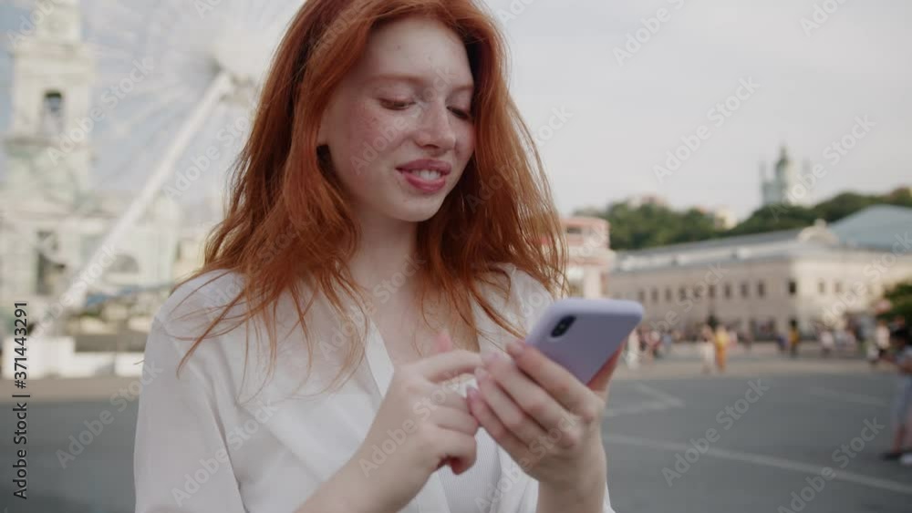 Ginger beautiful woman with earphones chatting online on smartphone while listening to music in earphones walking on street city center. People and technologies. Ginger beautiful woman with earphones chatting online on smartphone while listening to music in earphones walking on street city center. People and technologies.