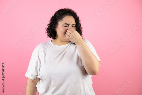 Young beautiful woman standing over isolated pink background smelling something stinky and disgusting, intolerable smell, holding breath with fingers on nose