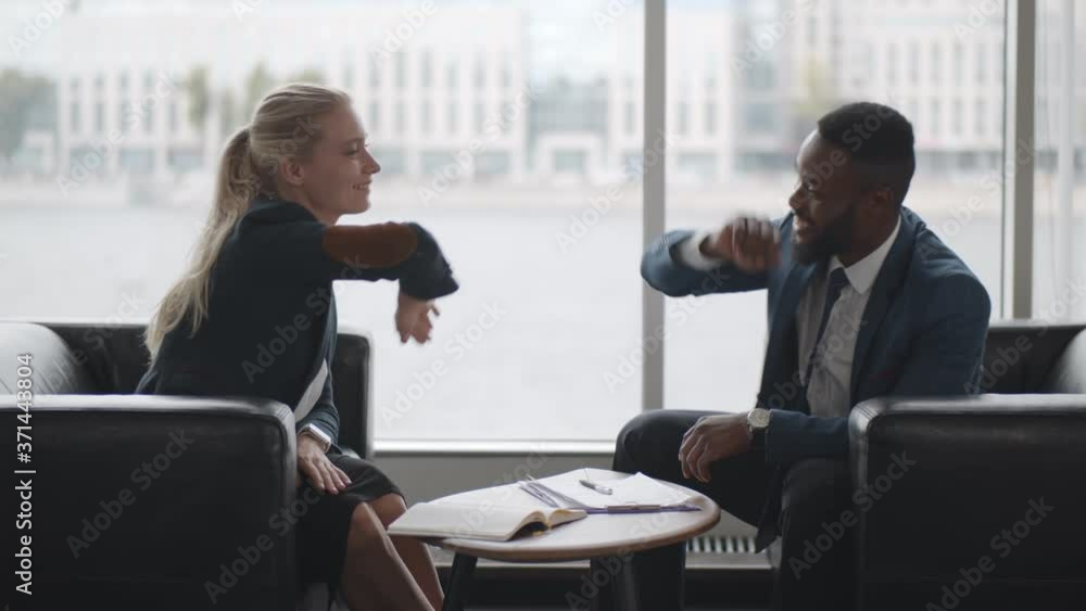 Side view of two multiethnic business colleagues sharing high five sitting on couch in office hall Side view of two multiethnic business colleagues sharing high five sitting on couch in office hall