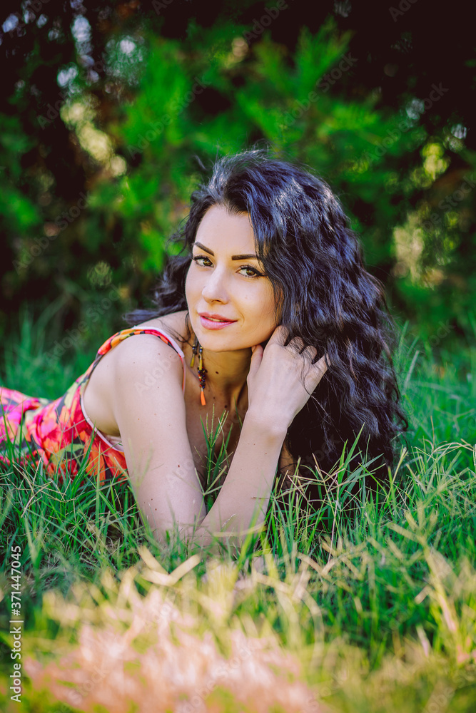 Beautiful curly-haired brunette with beautiful breasts in a colorful sundress lies on the grass Beautiful curly-haired brunette with beautiful breasts in a colorful sundress lies on the grass