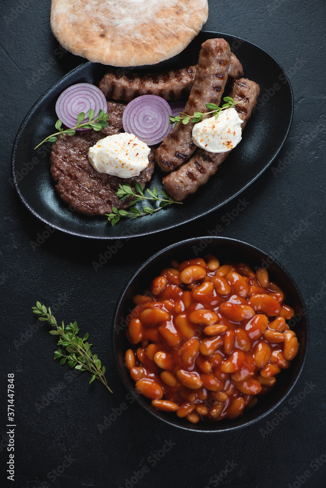 Grilled serbian pljeskavica and cevapi sausages with prebranac and pita, top view on a black stone background, vertical shot