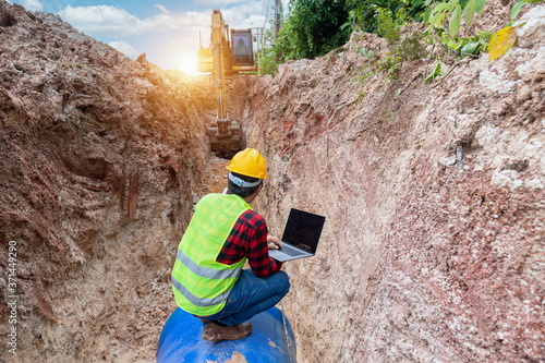 Fotografie Engineer wear safety uniform use laptop examining excavation Drainage Pipe and Large plumbing water system underground at construction site