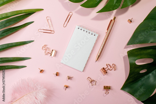 Flat lay of blank notepad, golden office supplies and palm leaves on pink background