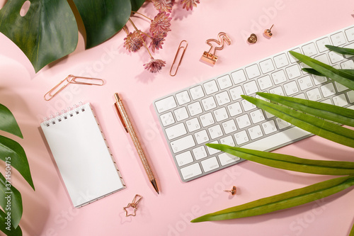 Flat lay of empty notepad, keyboard, golden office supplies and palm leaves on pink background