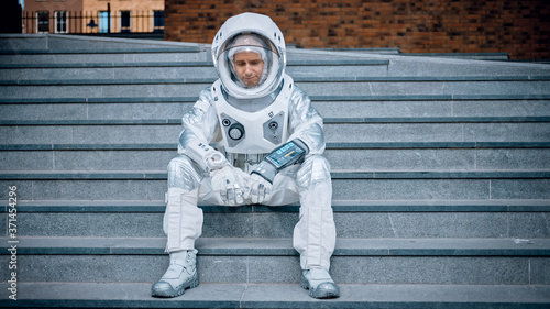 Sad Man in Spacesuit is Sitting on Concrete Stairs. Astronaut is Feeling Down and Looks Down. Emotionally Depressed Spaceman in White Futuristic Suit with Technological Panel on His Hand.