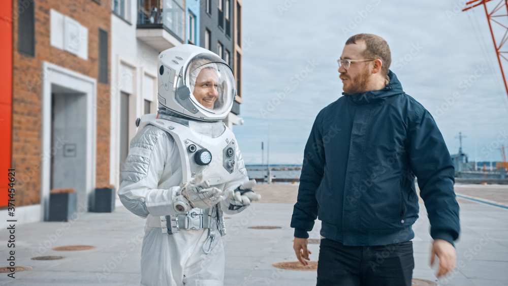 Ironic and Satirical Shot of Man in Spacesuit Walking on a Street with a Friend in Casual Clothes. Two Men Walk and Make a Pleasant Conversation. Man in Suit with Technological Panel on His Hand.