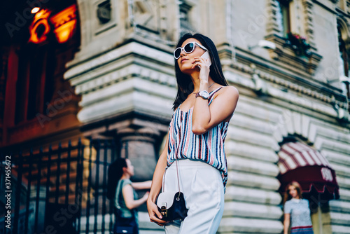 Serious female in sunglasses and trendy outfit talking on phone walking on city street. attractive young woman having mobile conversation using connection in roaming strolling during free time