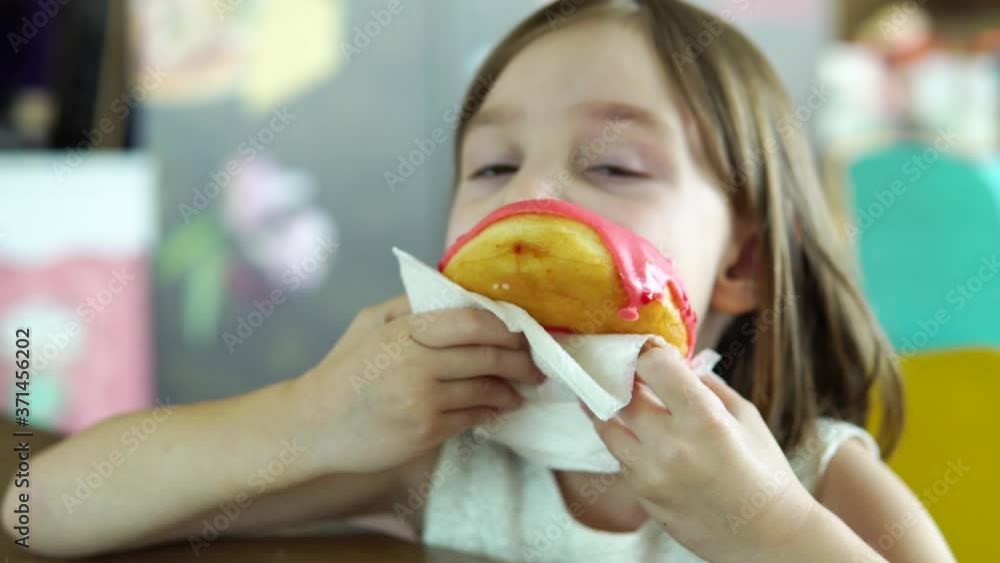 Little blonde girl funny eats a donut while sitting at a table at a food court or restaurant. Little blonde girl funny eats a donut while sitting at a table at a food court or restaurant.