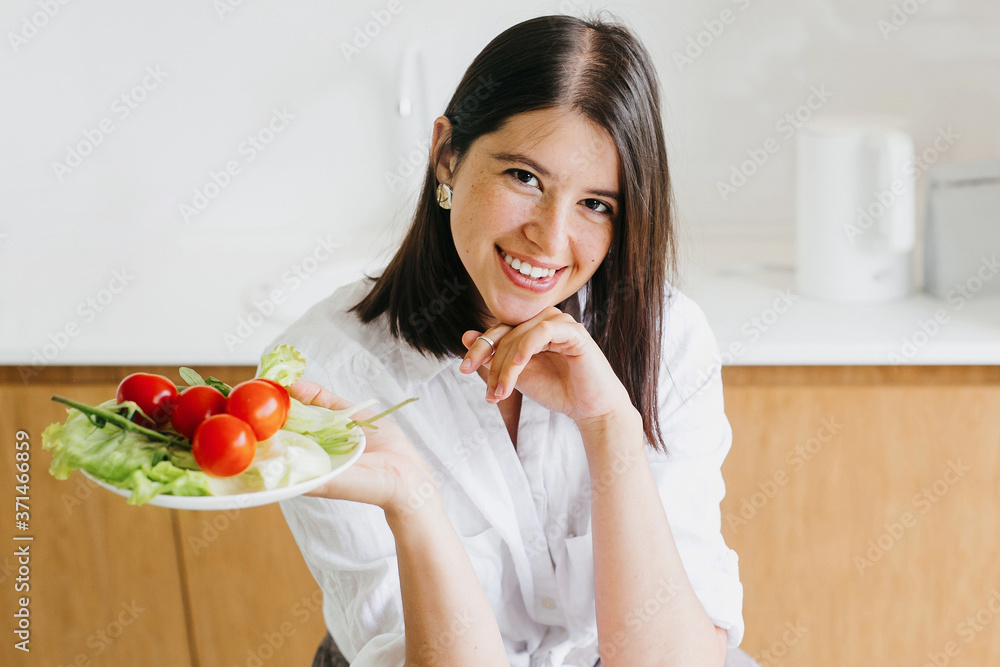 Young happy woman holding plate with green lettuce,arugula and cherry tomato on background of modern white kitchen. Healthy food concept. Home cooking, salad ingredients Young happy woman holding plate with green lettuce,arugula and cherry tomato on background of modern white kitchen. Healthy food concept. Home cooking, salad ingredients