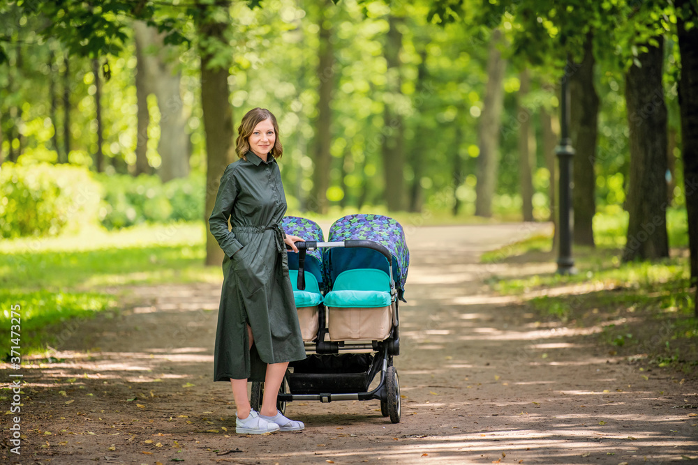 Happy young mom walks in the park in the summer with a stroller for twins. - obrazy, fototapety, plakaty 