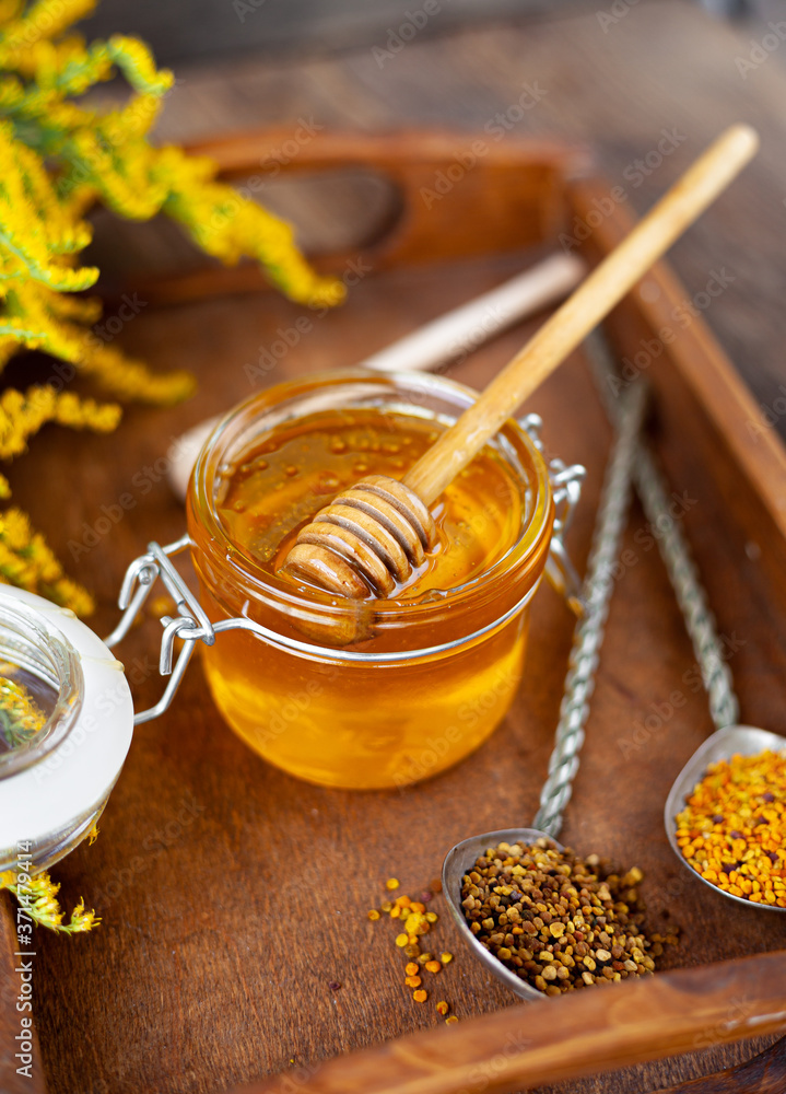 Honey in a glass jar with a wooden spoon. Still life on a wooden table with yellow flowers Goldenrod. Healthy sweet food. Close up with selective focus. Honey in a glass jar with a wooden spoon. Still life on a wooden table with yellow flowers Goldenrod. Healthy sweet food. Close up with selective focus.