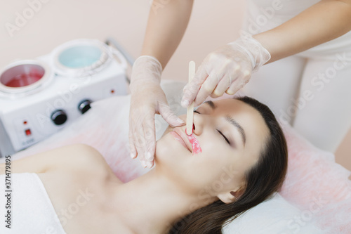 A master applies depilatory wax to the face of a beautiful young woman to remove unwanted hair. Sugaring. Depilation. Epilation. Beauty concept. Selective focus.