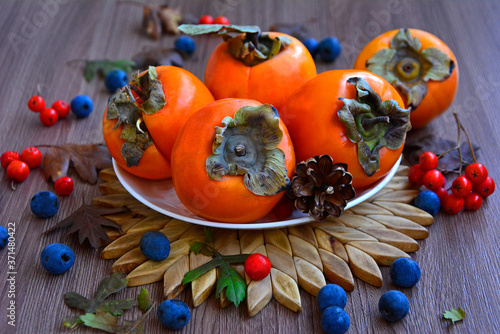 Ripe orange persimmon on a white plate, around the blue and red berries on a wooden background