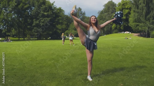 Beautiful cheerleader in uniform practicing split holding pompom standing outdoors photo