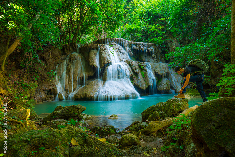 Naklejka premium PhotosSearch by image Young man with backpack standing near a waterfall in forest. Male hiker in the nature during rain