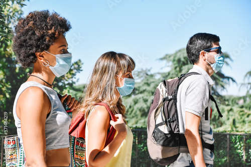 Three travel friends wearing face masks to avoid germs or pollution walk carrying their luggage. Tourism in pandemic times
