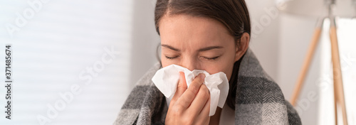 Sick woman feeling unwell staying home. Young girl with flu symptoms coughing in tissue covering nose when sneezing as COVID 19 prevention. Panoramic crop.