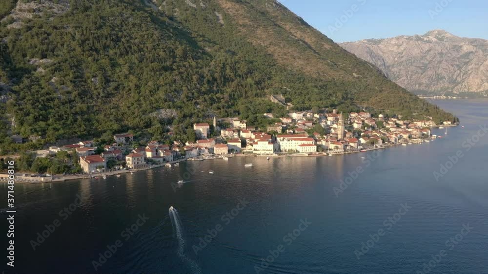 Flight over the Adriatic sea coast, small Montenegrin town Perast. We can see an ancient church, roofs of white stone houses and trees. It's sunset. Mountains in the background Flight over the Adriatic sea coast, small Montenegrin town Perast. We can see an ancient church, roofs of white stone houses and trees. It's sunset. Mountains in the background