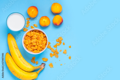 Fototapeta Naklejka Na Ścianę i Meble -  Breakfast cereals, bananas, apricots and a glass of milk on a bright blue background top view, flat lay. Fitness food concept. Breakfast with cornflakes, milk and fruit. Copy space.