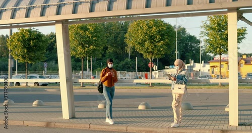 Muslims young pretty women in traditional hijabs standing outdoors and using gadgets. Arabic females in medical masks tapping and scrolling on mobile phone and tablet device. Arabs girls at bus stop. photo