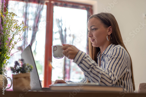 Young brunette woman drinking coffee and looking in her laptop computer.
