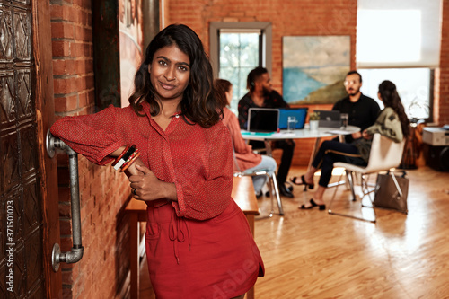 portrait young ethnic woman standing and leaning on sliding door, coworkers gathered around desk in background  photo