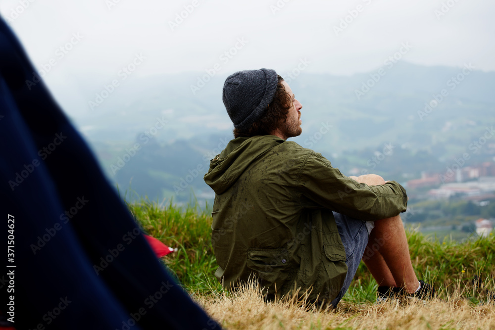 Fototapeta premium Young hike hipster man sitting at his campsite on the top of hill while enjoying the nature in rainy weather, young man relaxing on the grass enjoying outdoors view and relaxing after hike way