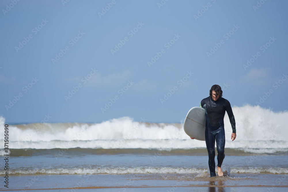 Young surfer sport man carrying his surfing board and going out the sea while on vacation during a sunny day, with an intense blue sky and big waves break on background Young surfer sport man carrying his surfing board and going out the sea while on vacation during a sunny day, with an intense blue sky and big waves break on background