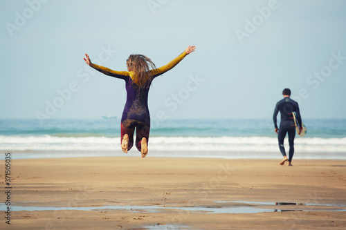 Photography Young surfer girl jump having fun while her friend walk to the ocean ready to su