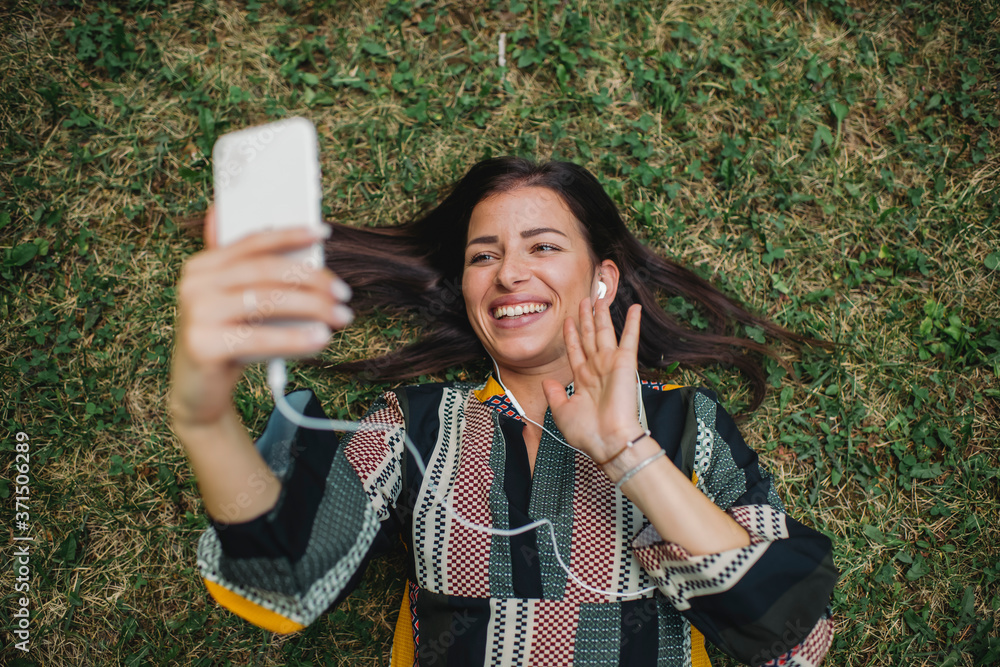 Fototapeta premium Young Caucasian woman lying down in the grass and taking selfie