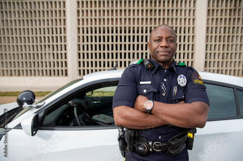 Portrait of Police officer Standing in street in front of squad car looking towards camera with arms behind his back 