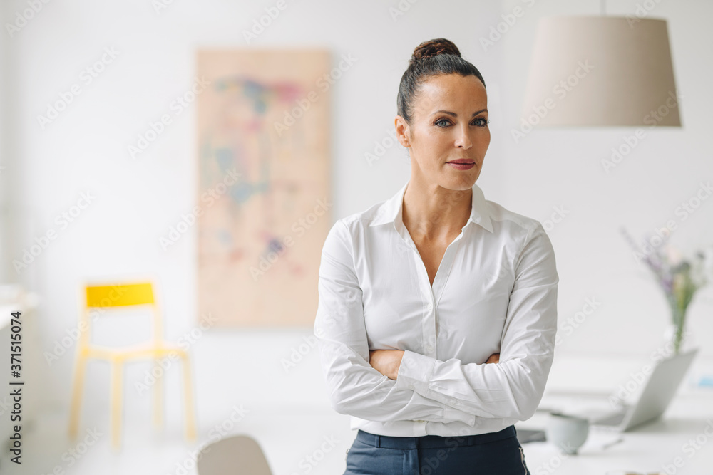 Female professional with arms crossed standing against wall in home office Female professional with arms crossed standing against wall in home office